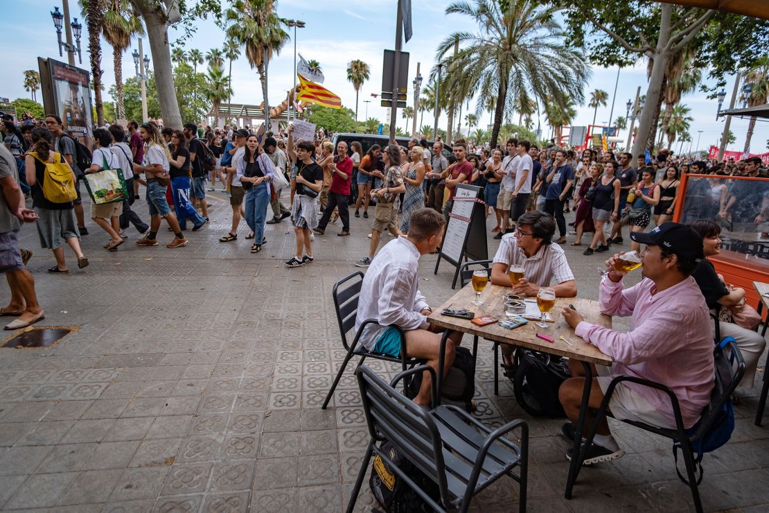 Ljudje pijejo pivo pred gostinskim lokalom, medtem ko mimo potekajo demonstracije proti turizmu v Barceloni julija 2024.